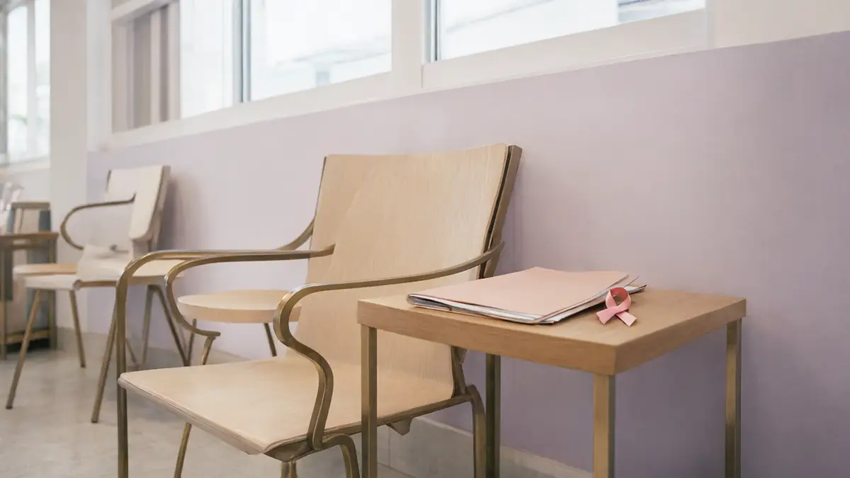 Sala de espera clara e vazia, com cadeiras de madeira, pasta fechada e um laço rosa sobre a mesa lateral, com uma janela grande ao fundo